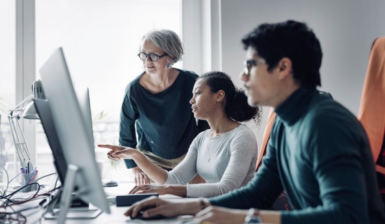 Three people are in an office, collaboratively working. One is pointing at a computer screen, while another leans in to look at the same screen.