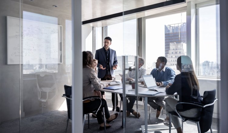 A group of businesspeople, dressed in smart business clothes, are gathered around a table in a glass-walled office. They are mid discussion, with one person standing and gesticulating with their hands, and the others sat listening attentively.