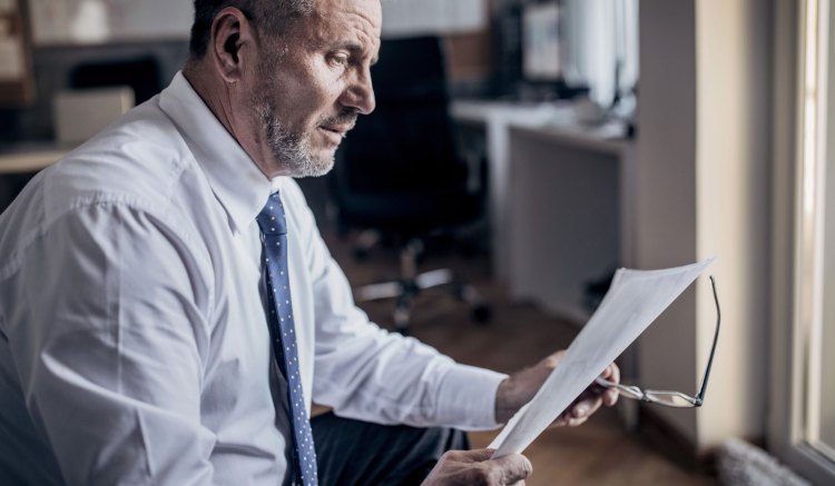 A man in a white shirt and blue tie is sat in an office. He is holding a pair of spectacles in one hand as he reads a piece of paper he is holding in the other. He looks concerned.