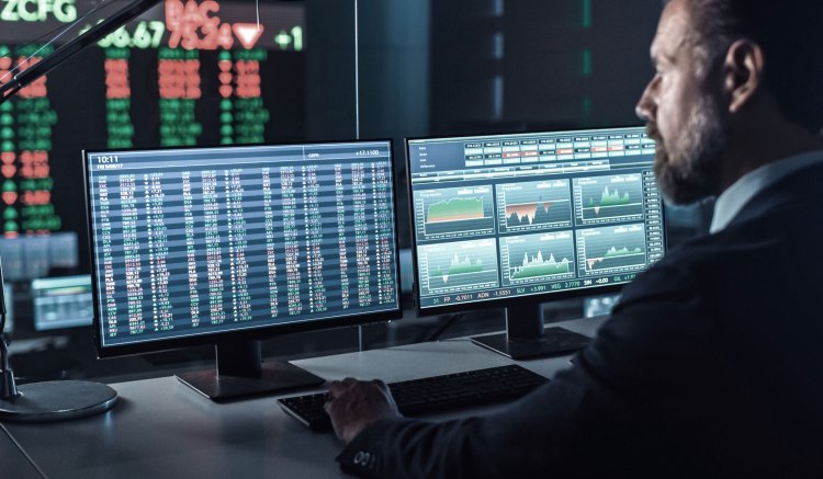 A person in business attire is sat in a dark office, using two computer screens to review market and trading data. On the wall in front of them is a large LED screen showing more trading and market information.