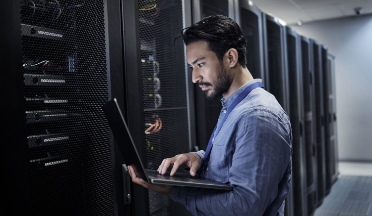 A bearded man wearing a blue shirt is stood in front of a row of server stacks in an office environment. He is holding an open laptop and typing on the keyboard.