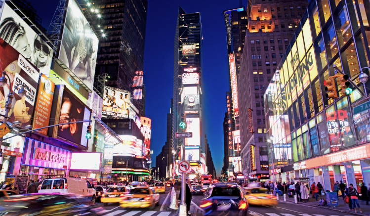 A picture of times square, New York, at night. It is brightly lit from digital advertising billboards, and the streets are crowded with people and yellow taxis.