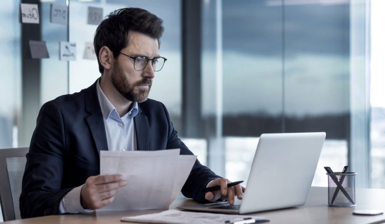 A man in a dark suit and a light blue shirt is sat in an office. He holds several sheets of paper in his right hand and is looking at a laptop on the desk in front of him. He has a beard, dark hair, and glasses.