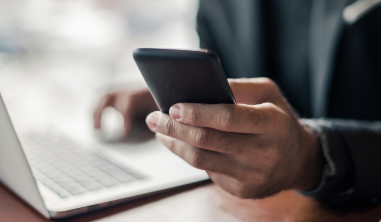 This image shows a person, wearing a dark suit, sat at a desk. It is shot so that only the person's hands are in frame - they are holding a smartphone in one hand, and operating a silver laptop with the other hand