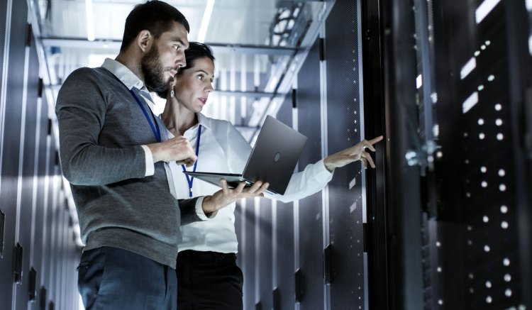 The image is of two people wearing business clothing stood in a data center, pointing and looking at a server, perhaps discussing data completeness strategy.