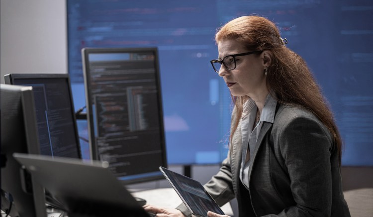 A female compliance officer sits in front of multiple computer monitors.
